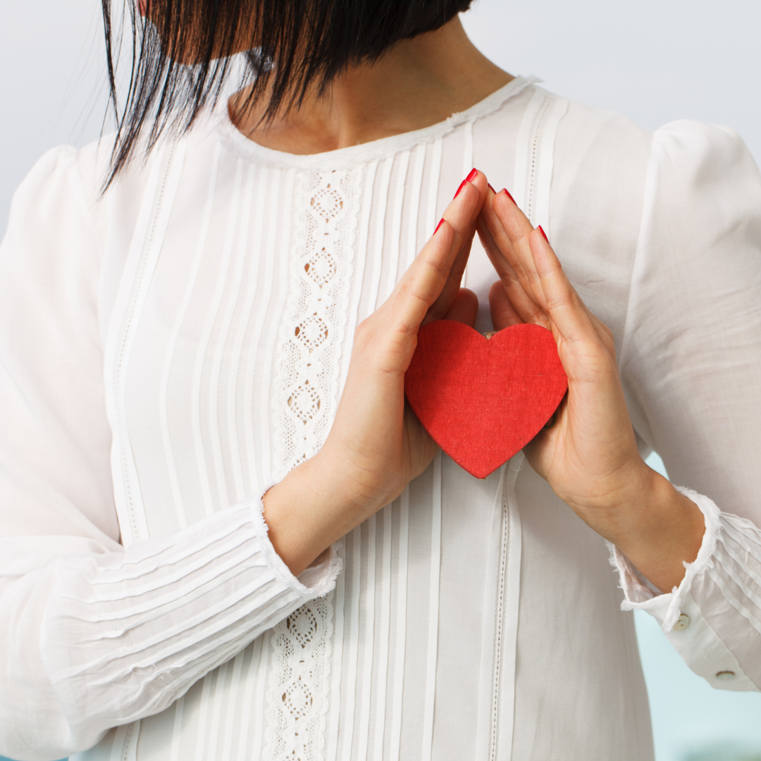 woman wearing a white blouse while holding a red heart shape in her hands close to her chest.
