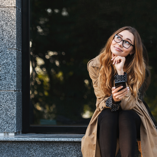 a woman wearing eyeglasses and a brown coat sitting a window ledge with her phone in her hand and looking up, smiling.
