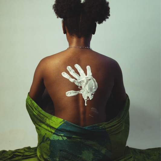 Black woman wrapped in a green blanket sitting with her back exposing a large white handprint on her back.