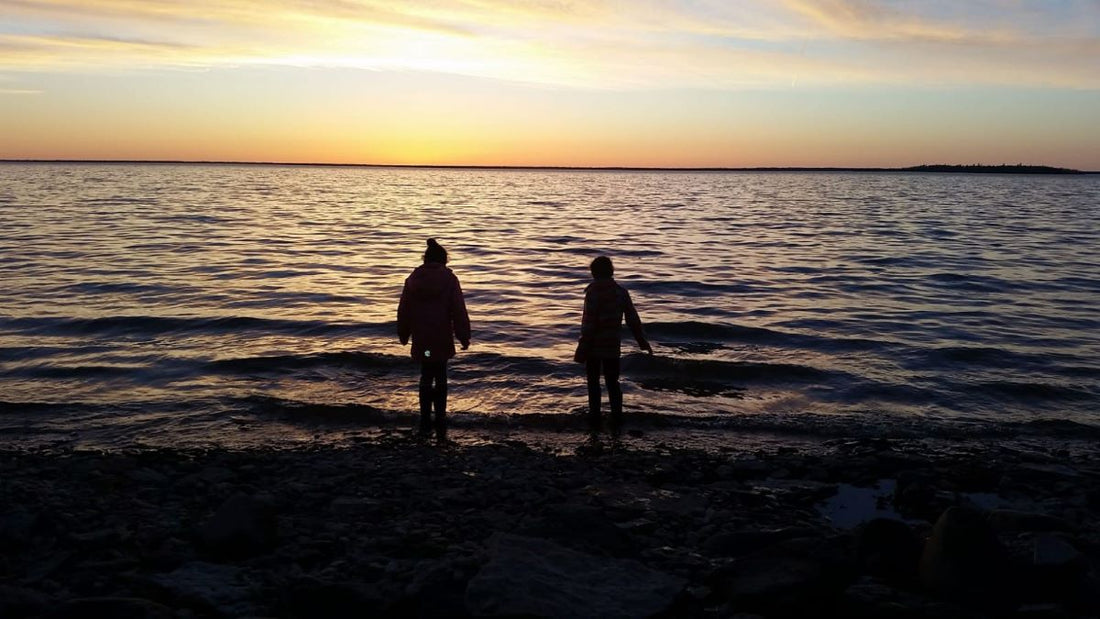 silhouette of two children along the lake shore as the sun sets in the background.