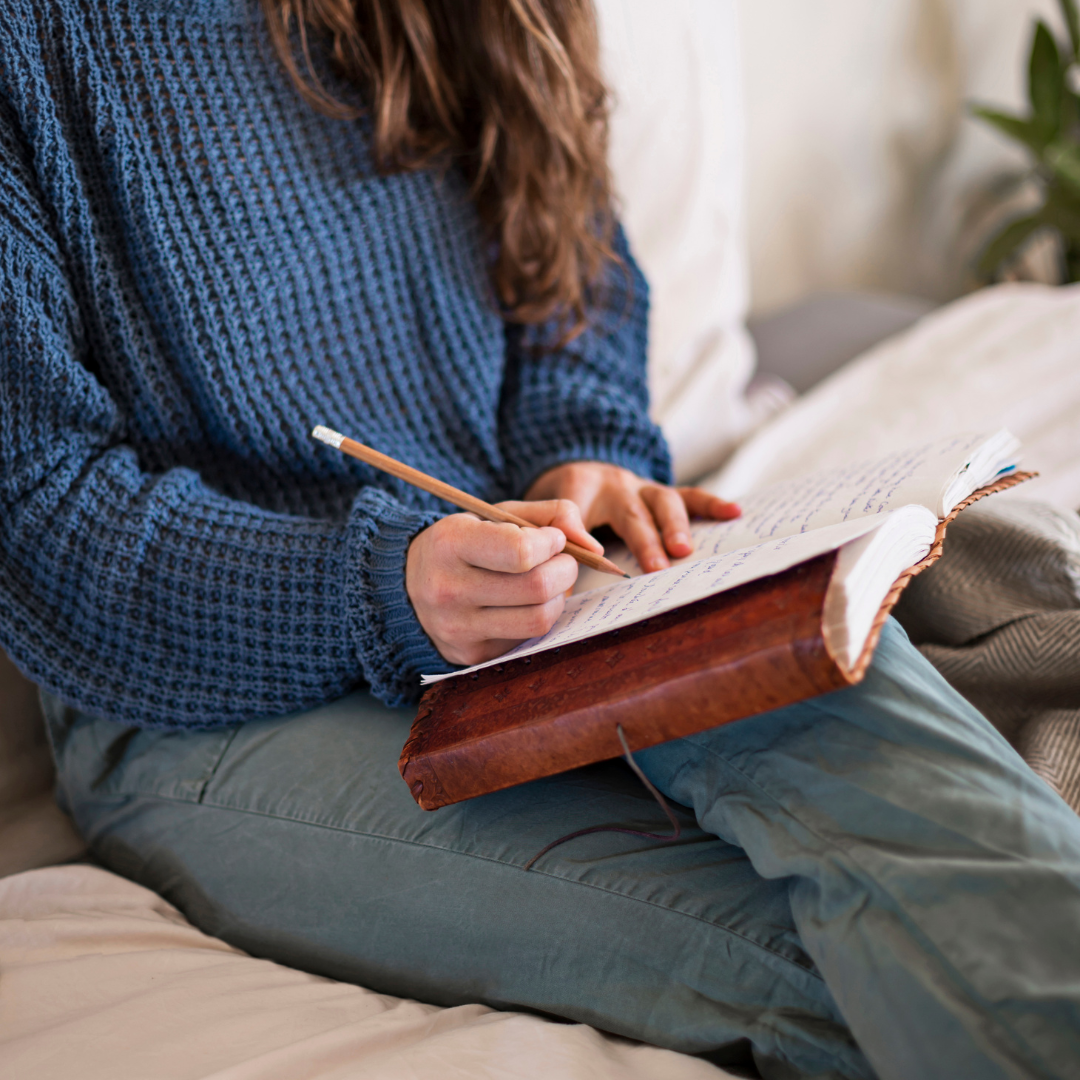 A photograph of a woman wearing a blue sweater and blue pants writing in a journal with a brown leather cover.