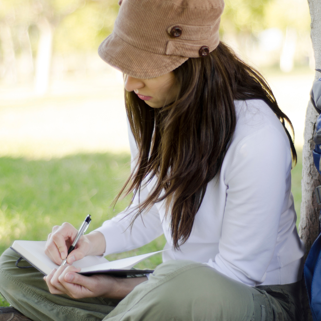 A woman wearing green pants and a white t-shirt is sitting on the grass and writing in a journal.