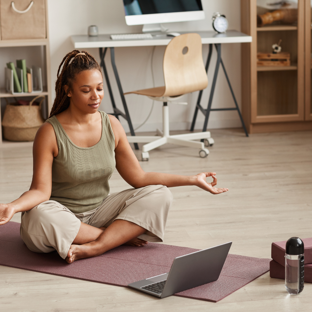 A photograph of a woman sitting on a yoga mat and holding a laptop.