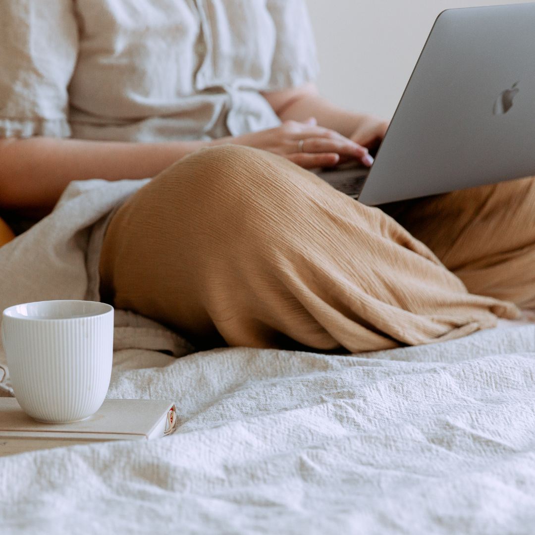 A person sitting on a bed holding a laptop in their lap. There is a coffee cup and a journal beside them.
