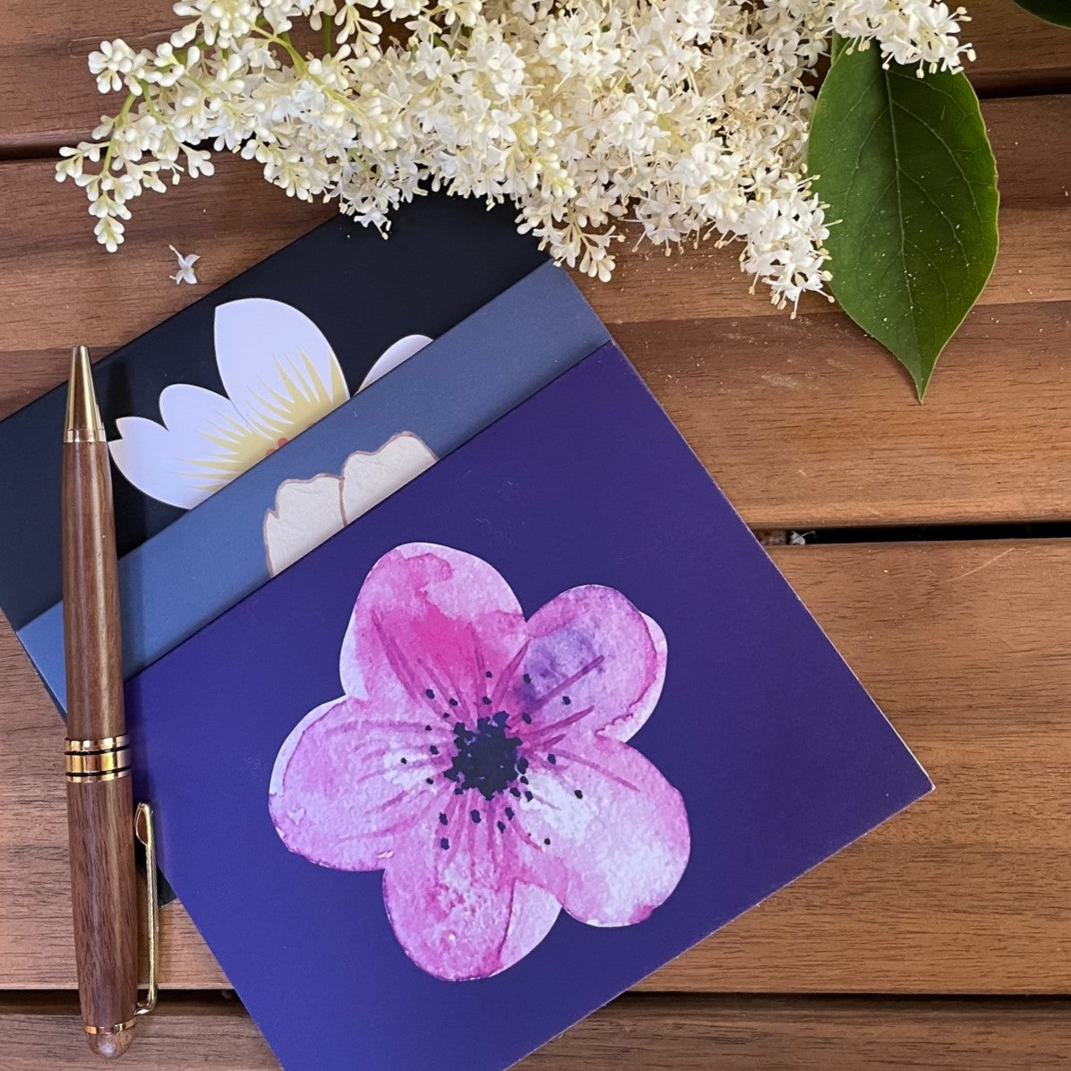 Hibiscus greeting and note cards, pack of six, black card with white graphic design flower, grey-blue card with yellow graphic design flower, navy blue card with graphic design flower, three postcards and brown pen laying on a wood background with a white flower.